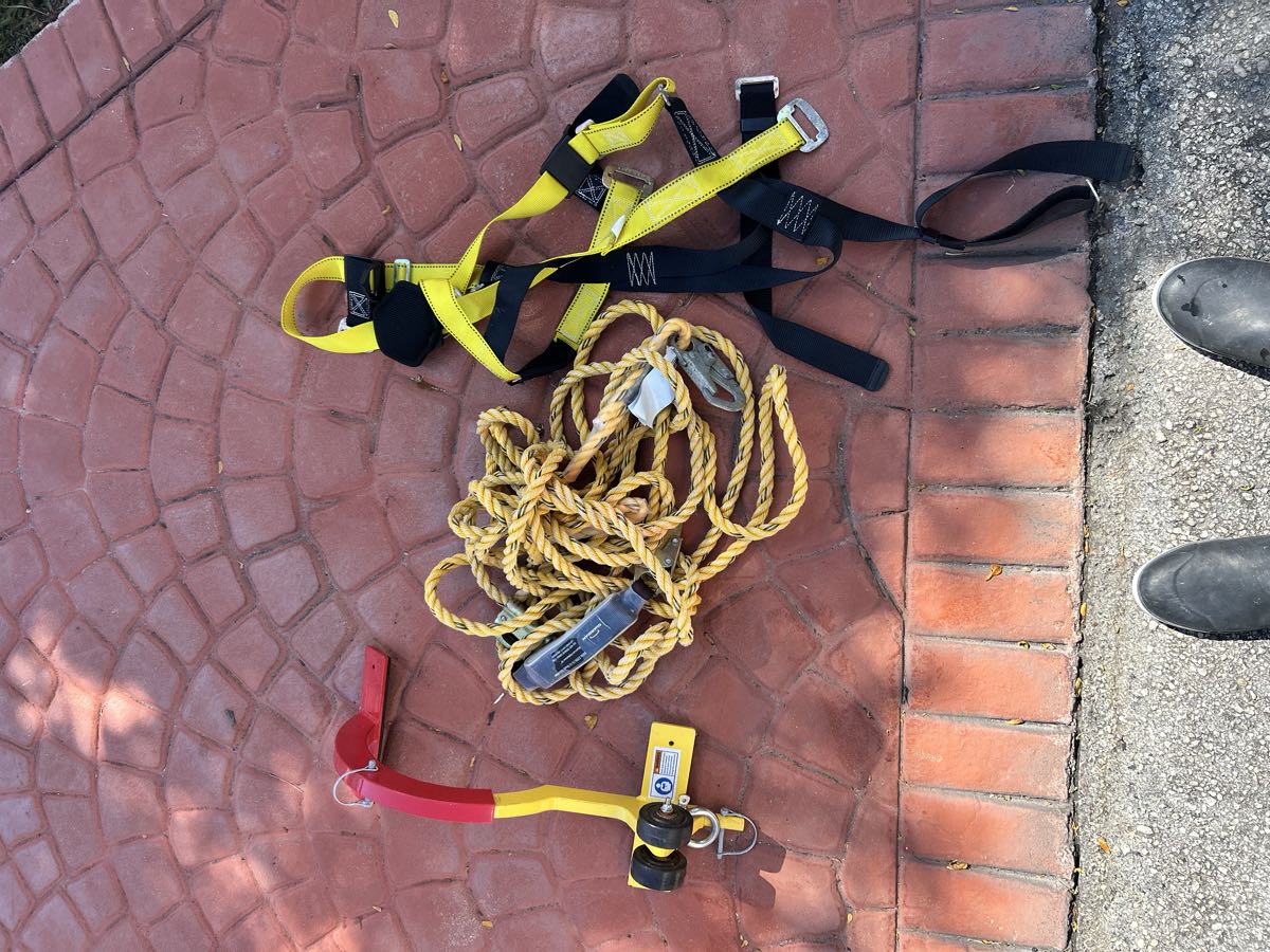 Roof safety gear laid out on pavers: a yellow full-body harness, a lifeline rope and a roof anchor